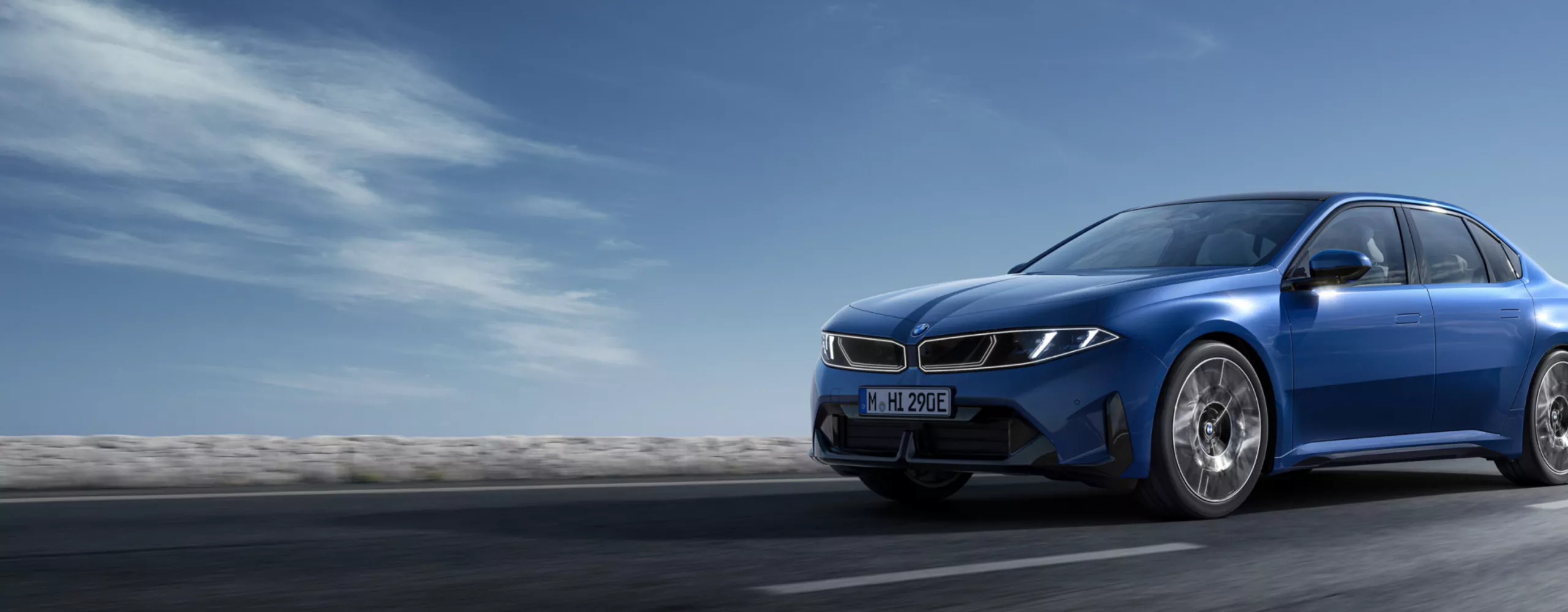 A blue sports sedan is captured in motion on an empty highway under a dramatic blue sky. The car is positioned prominently in the foreground, emphasizing its sleek design and dynamic presence. The setting is outdoors with a wide, open road and minimal traffic, creating a sense of freedom and speed. The overall mood is energetic and modern, with cool tones dominating the scene. A numeric license plate is visible on the vehicle.