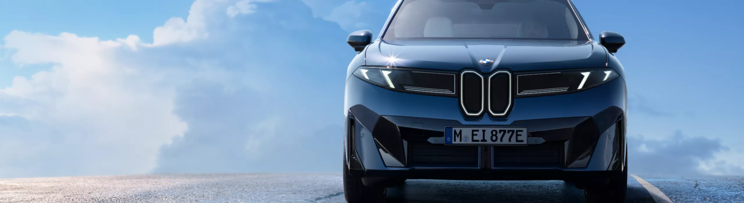 A BMW iX3 is driving on a wet, elevated road in front of a clear blue sky with few clouds