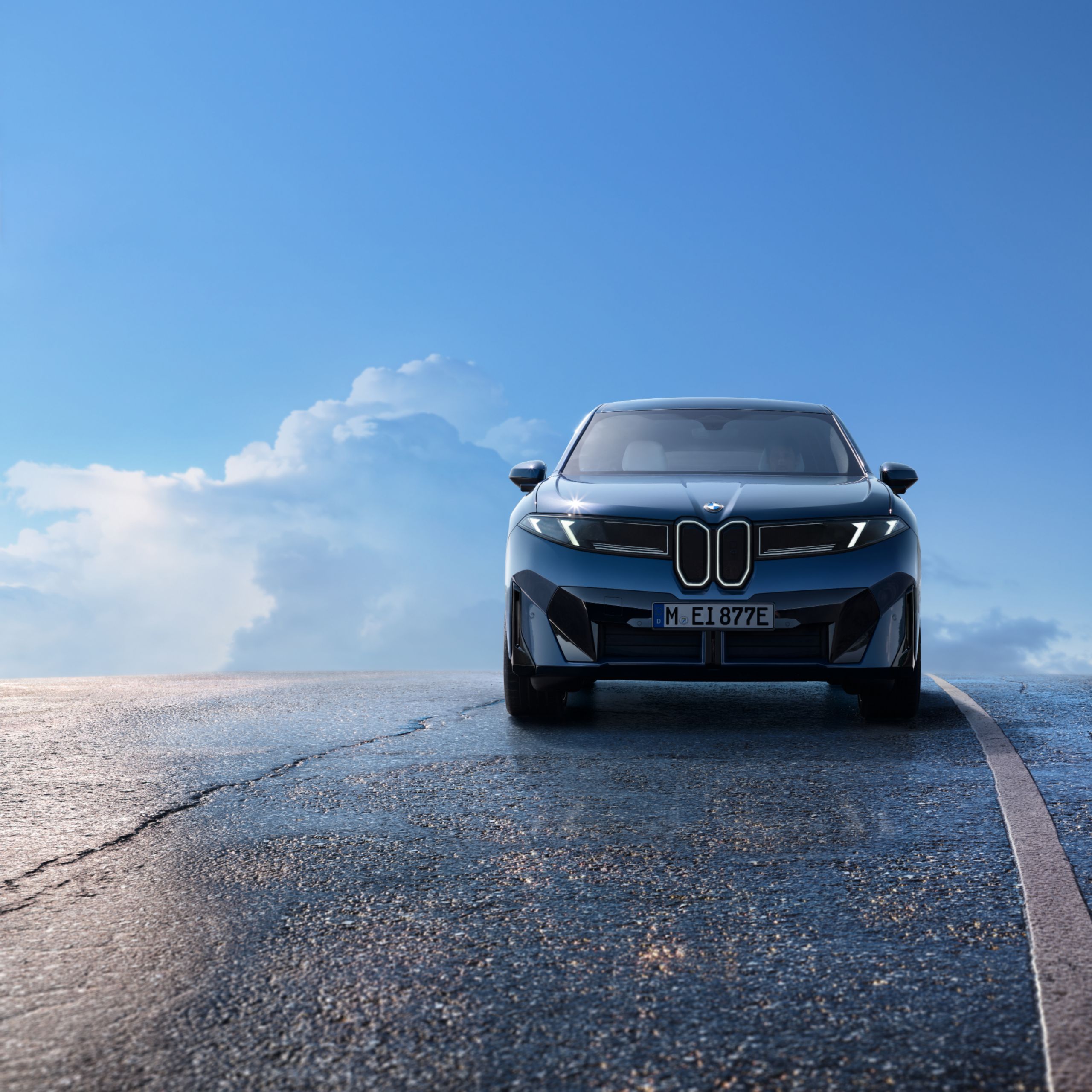 A BMW iX3 is driving on a wet, elevated road in front of a clear blue sky with few clouds