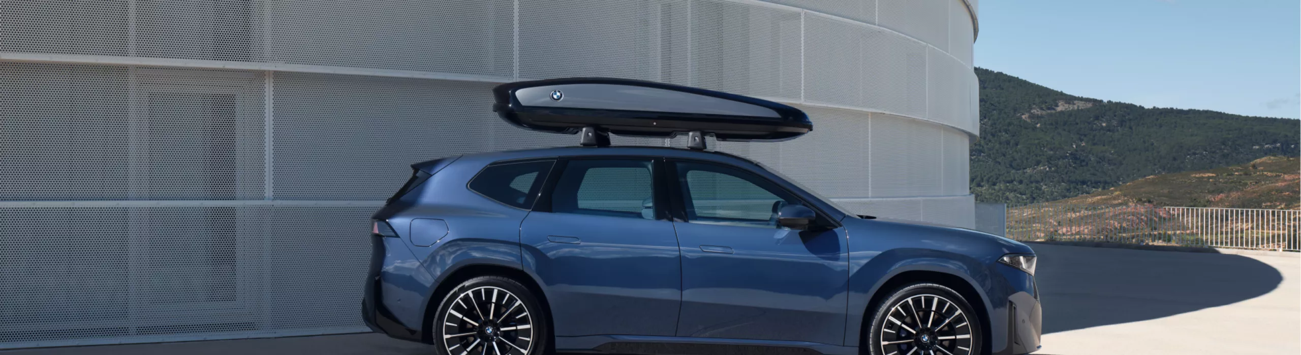 En BMW iX3 med takstativ er parkert på et parkeringsdekk foran en imponerende fjellutsikt