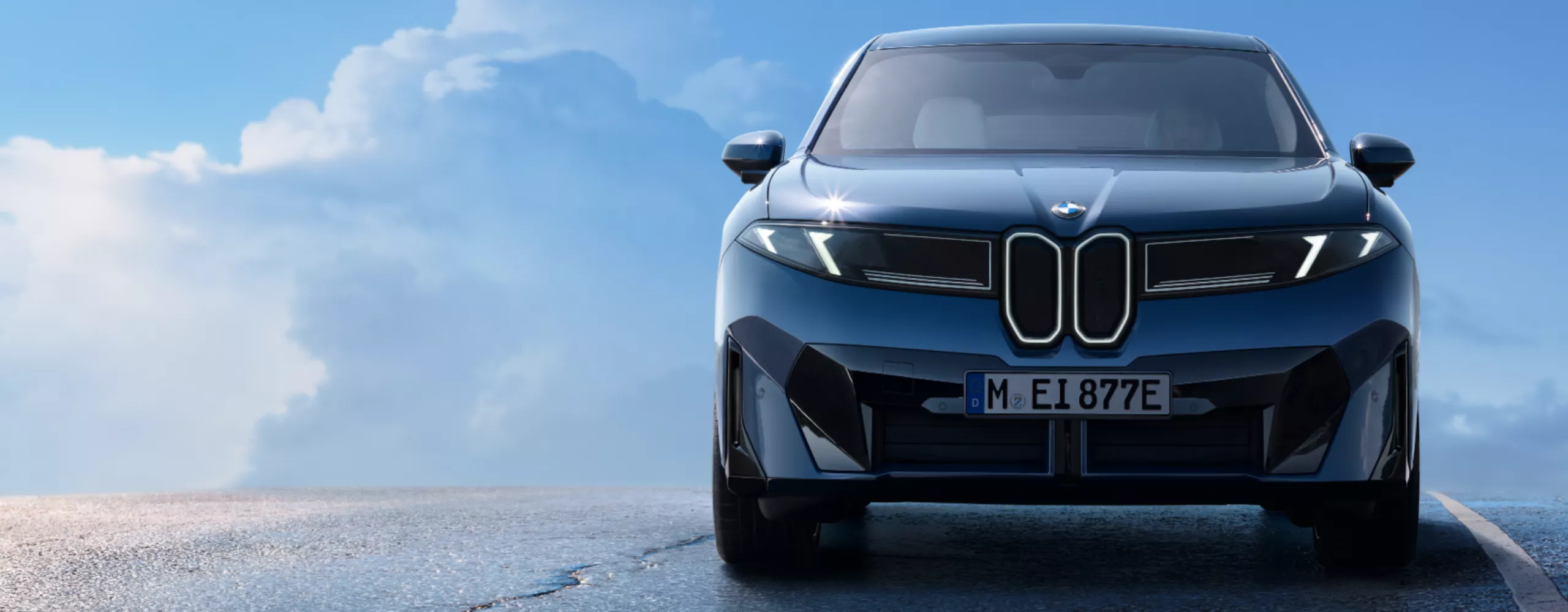 BMW iX3 vozi po mokrom, povišenom putu ispred vedrog plavog neba sa nekoliko oblaka.