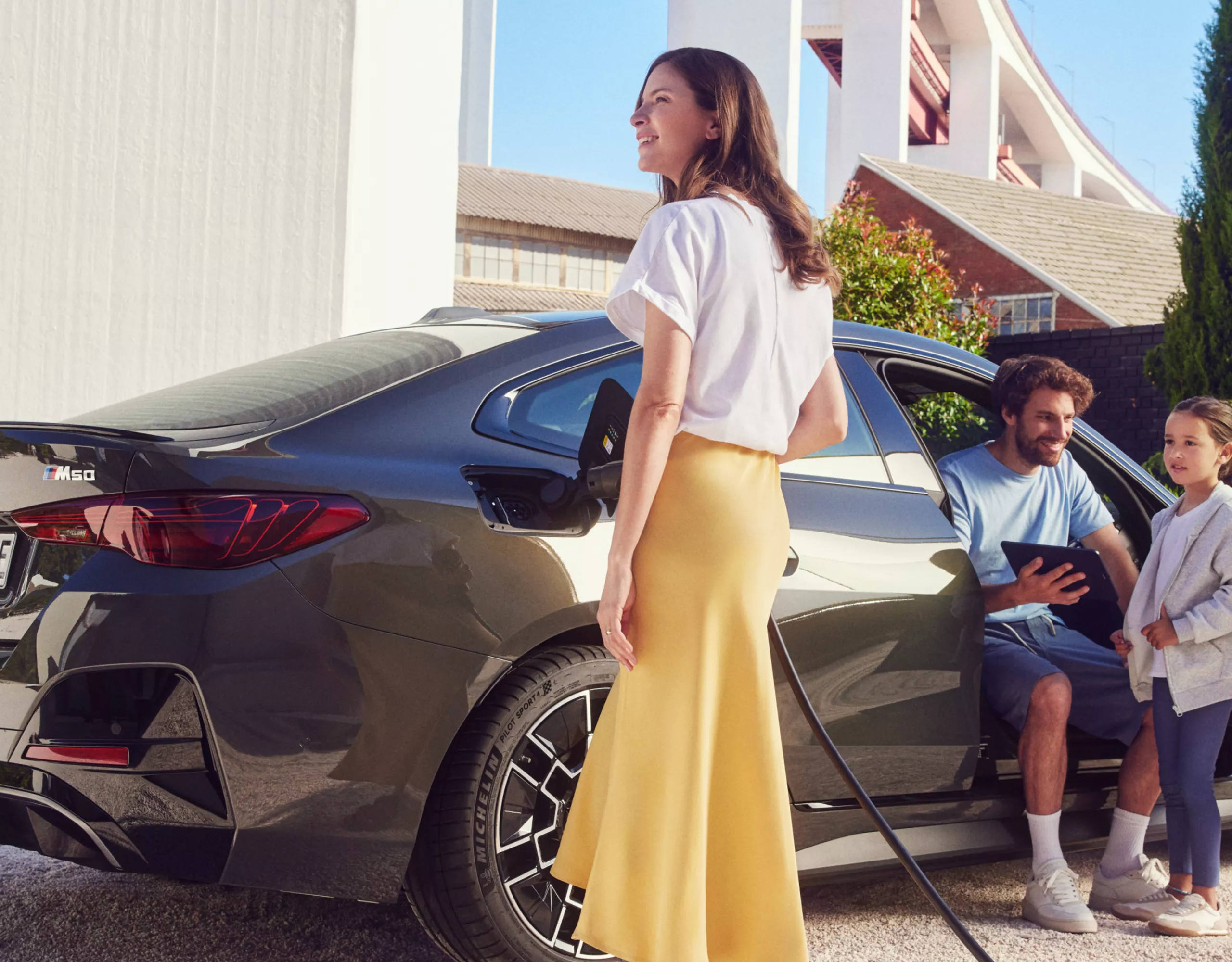 Eine Frau schließt ein Ladekabel an einen BMW i4 M50 xDrive an, Mann und Kind sind im Bild, im Hintergrund ist ein Haus