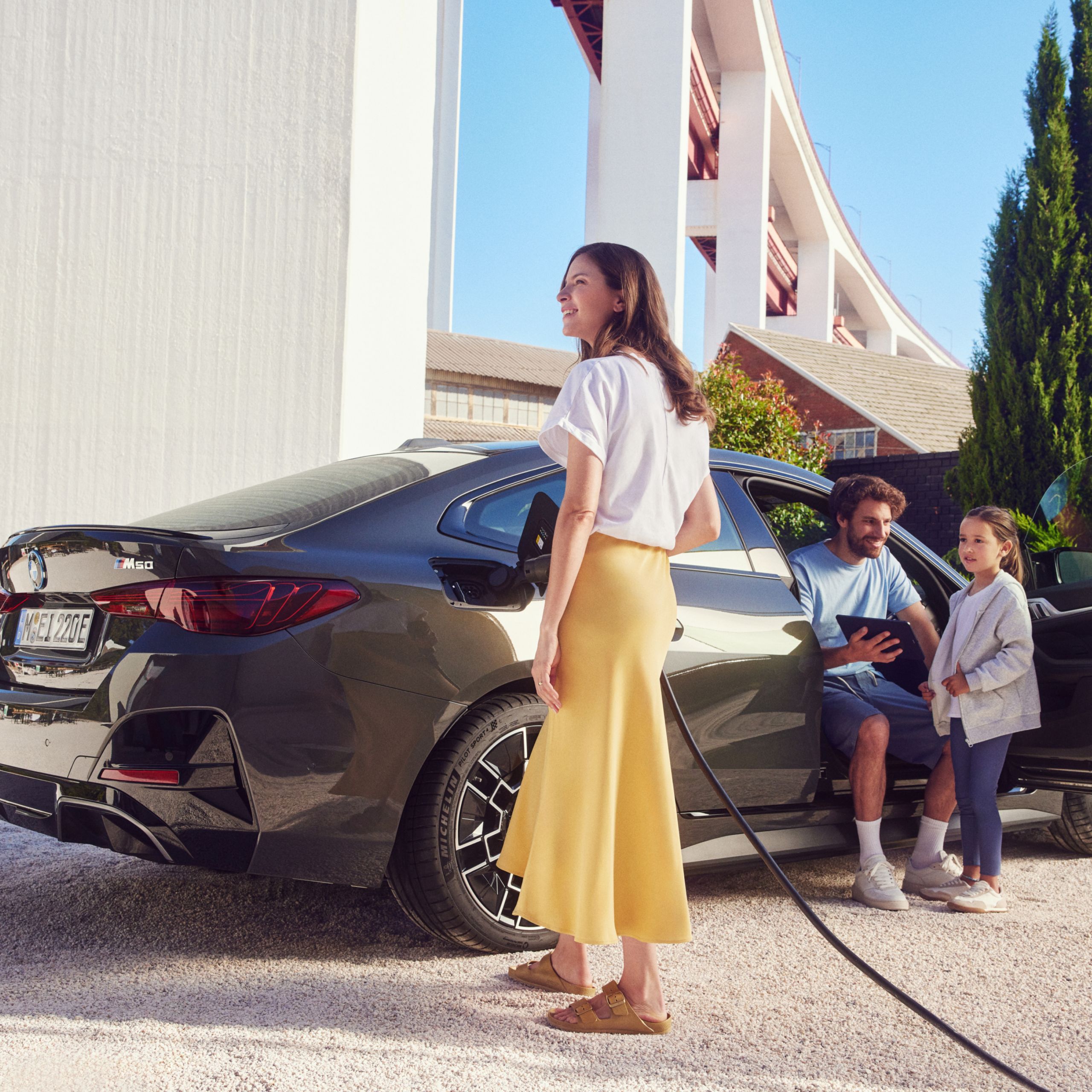 Une femme connecte un câble de recharge à une BMW i4 M50 xDrive, un homme et un enfant sont sur la photo, il y a une maison en arrière-plan
