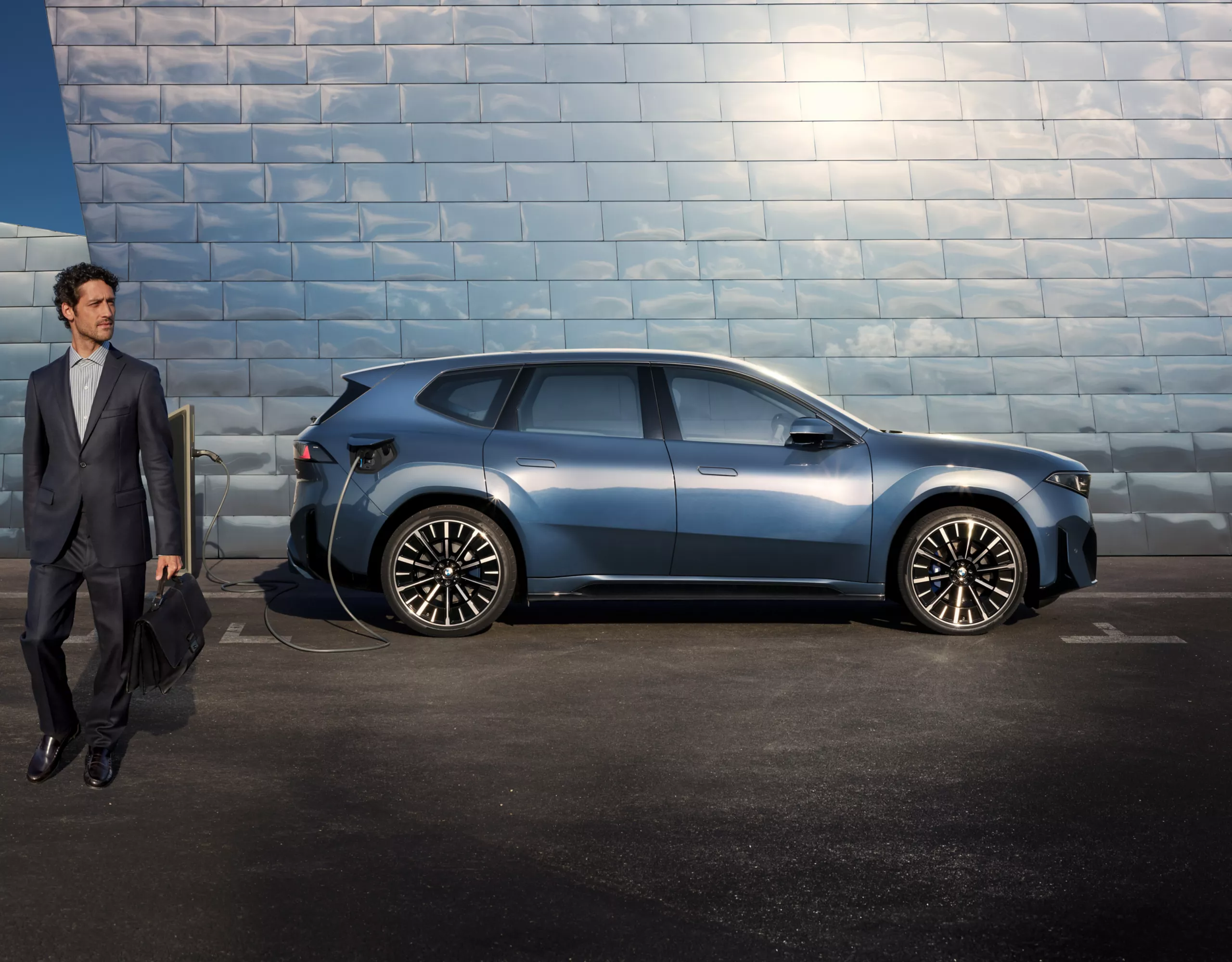 Un homme d'affaires s'éloigne d'une voiture de société BMW iX3 bleue garée devant la façade d'un bâtiment moderne et en métal