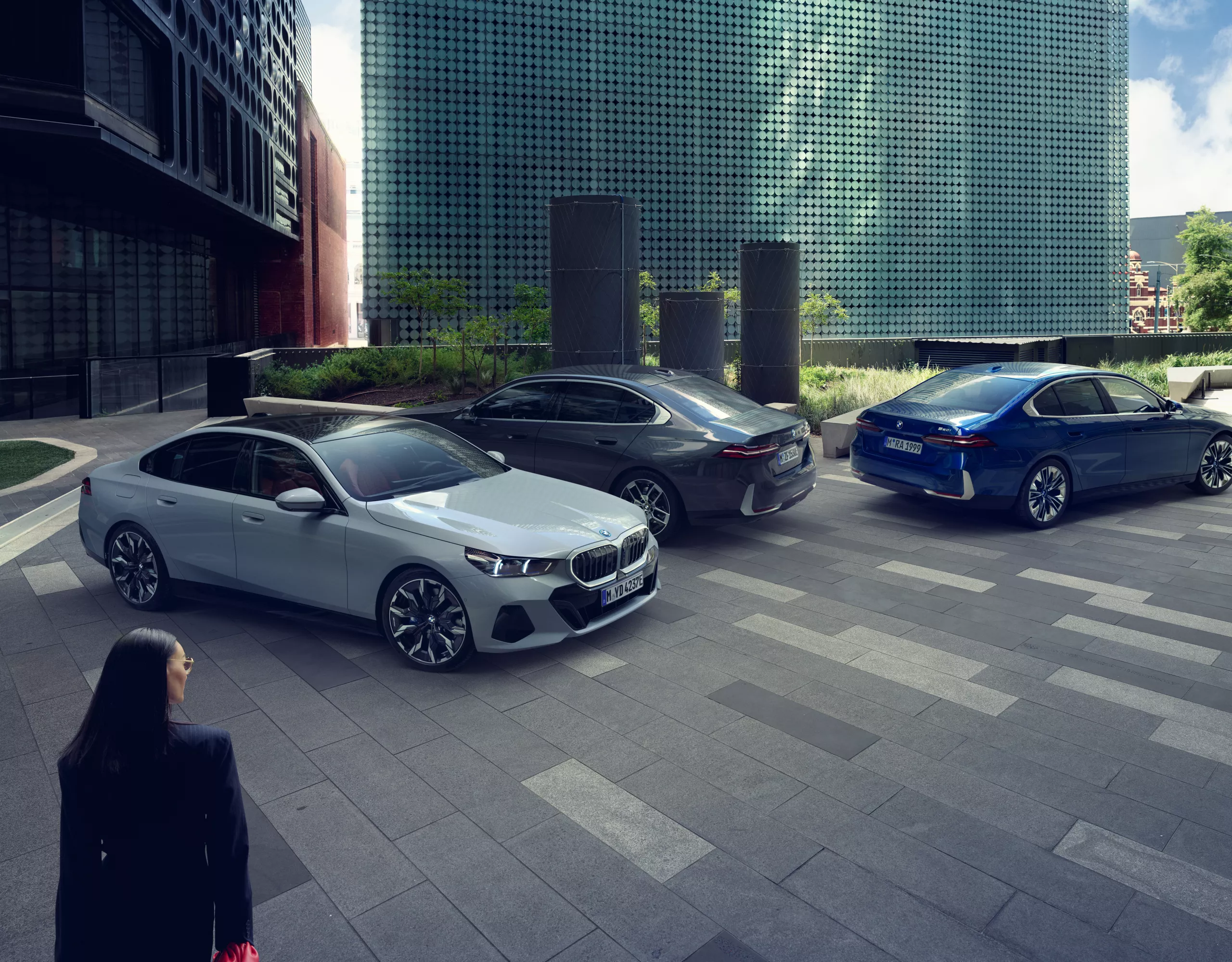 Trois modèles BMW sont garés sur une place moderne dans une ville, entourés de bâtiments avec beaucoup de verre et une personne au premier plan qui s'approche des véhicules