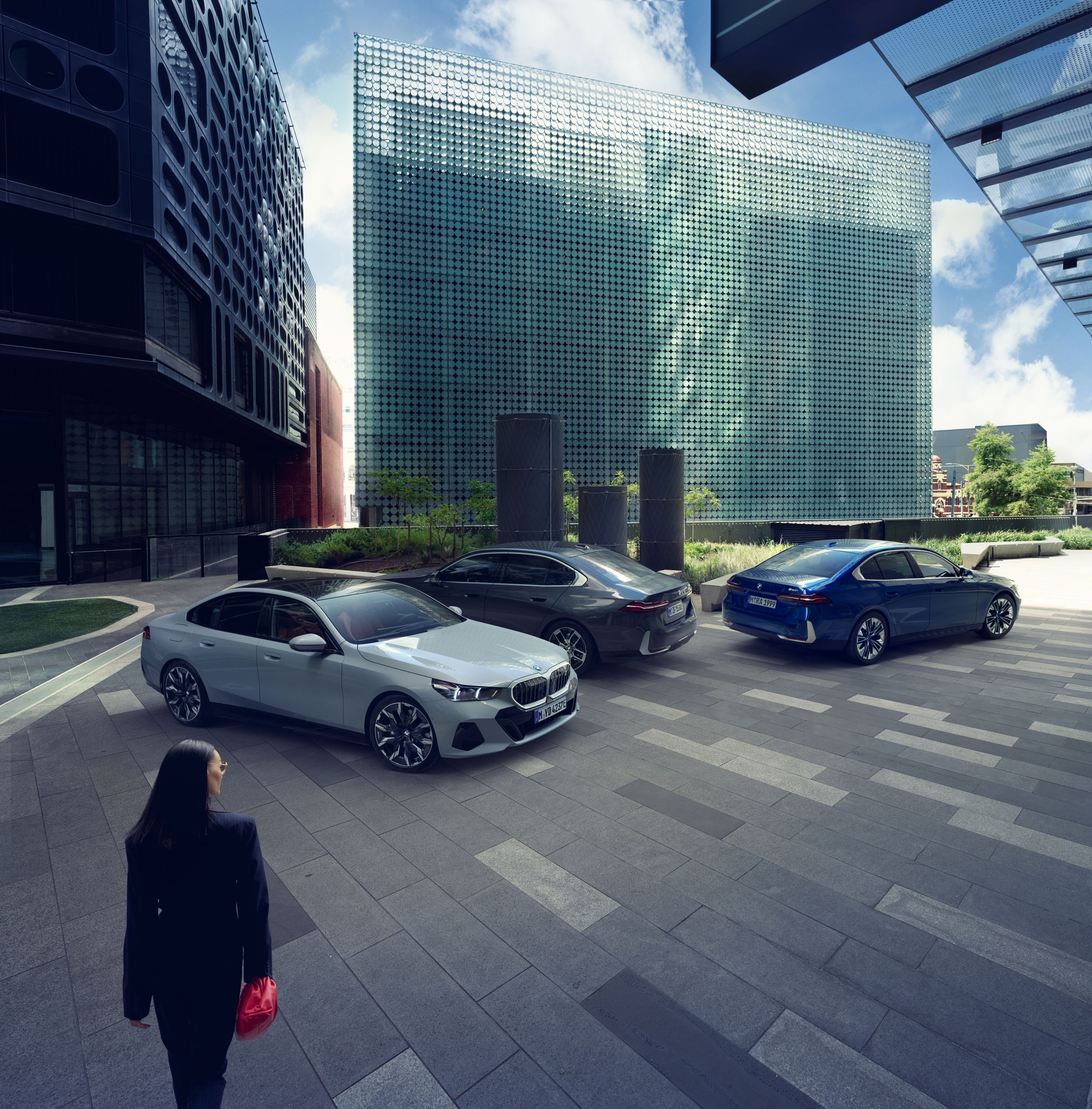 Trei modele BMW parcate într-o piață urbană modernă, înconjurată de clădiri din sticlă, cu o persoană care se îndreaptă spre automobilele din prim plan