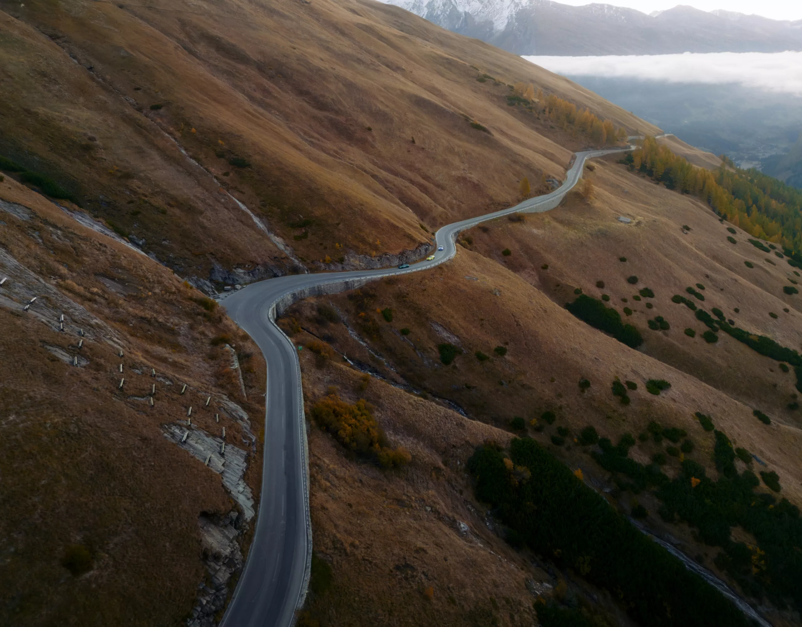 Kurvenreiche Bergstraße in einer herbstlich braunen Berglandschaft, im Hintergrund schneebedeckte Gipfel und Nebel. Drei BMW Fahrzeuge fahren die Straße entlang