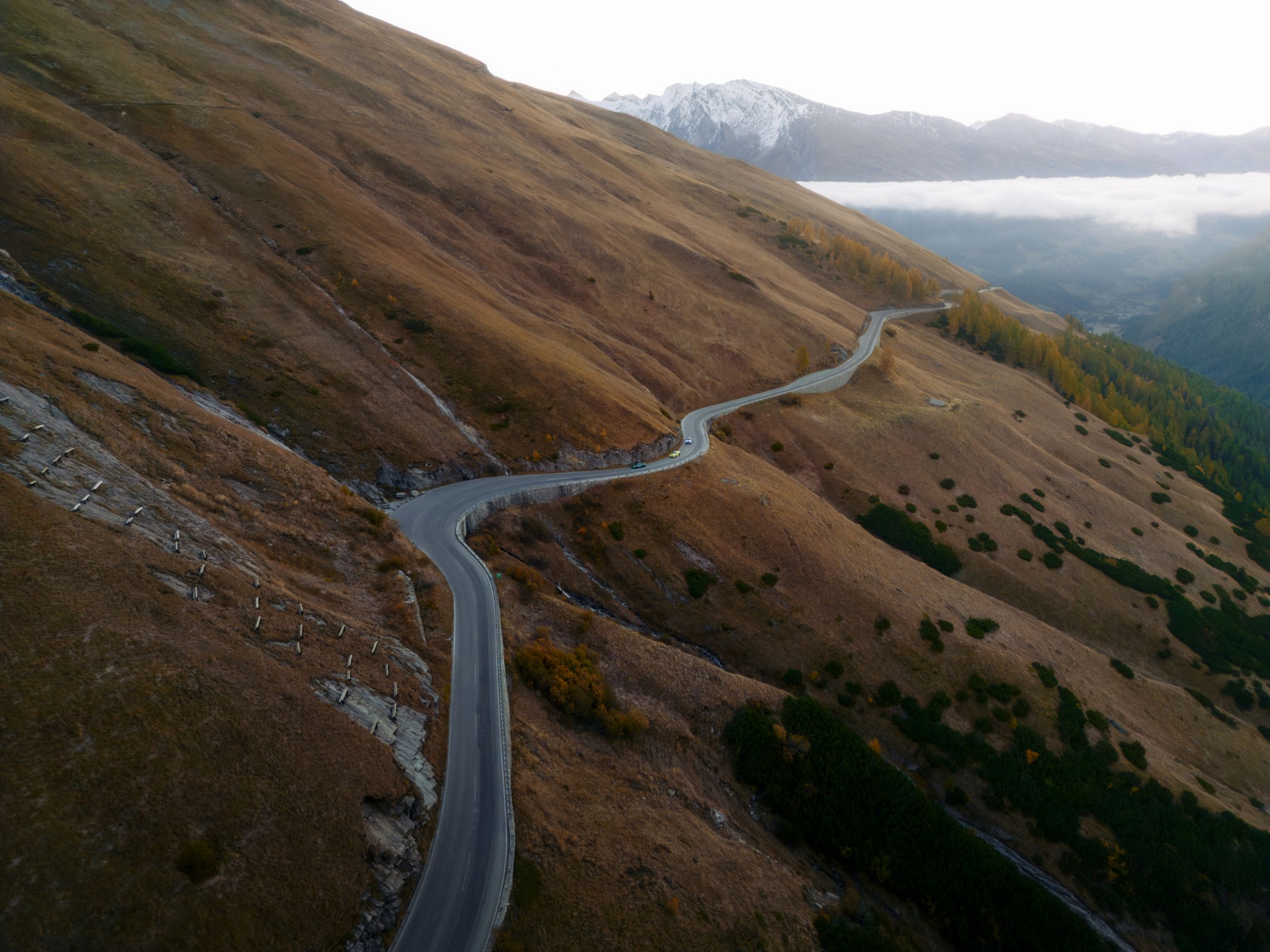Kurvenreiche Bergstraße in einer herbstlich braunen Berglandschaft, im Hintergrund schneebedeckte Gipfel und Nebel. Drei BMW Fahrzeuge fahren die Straße entlang