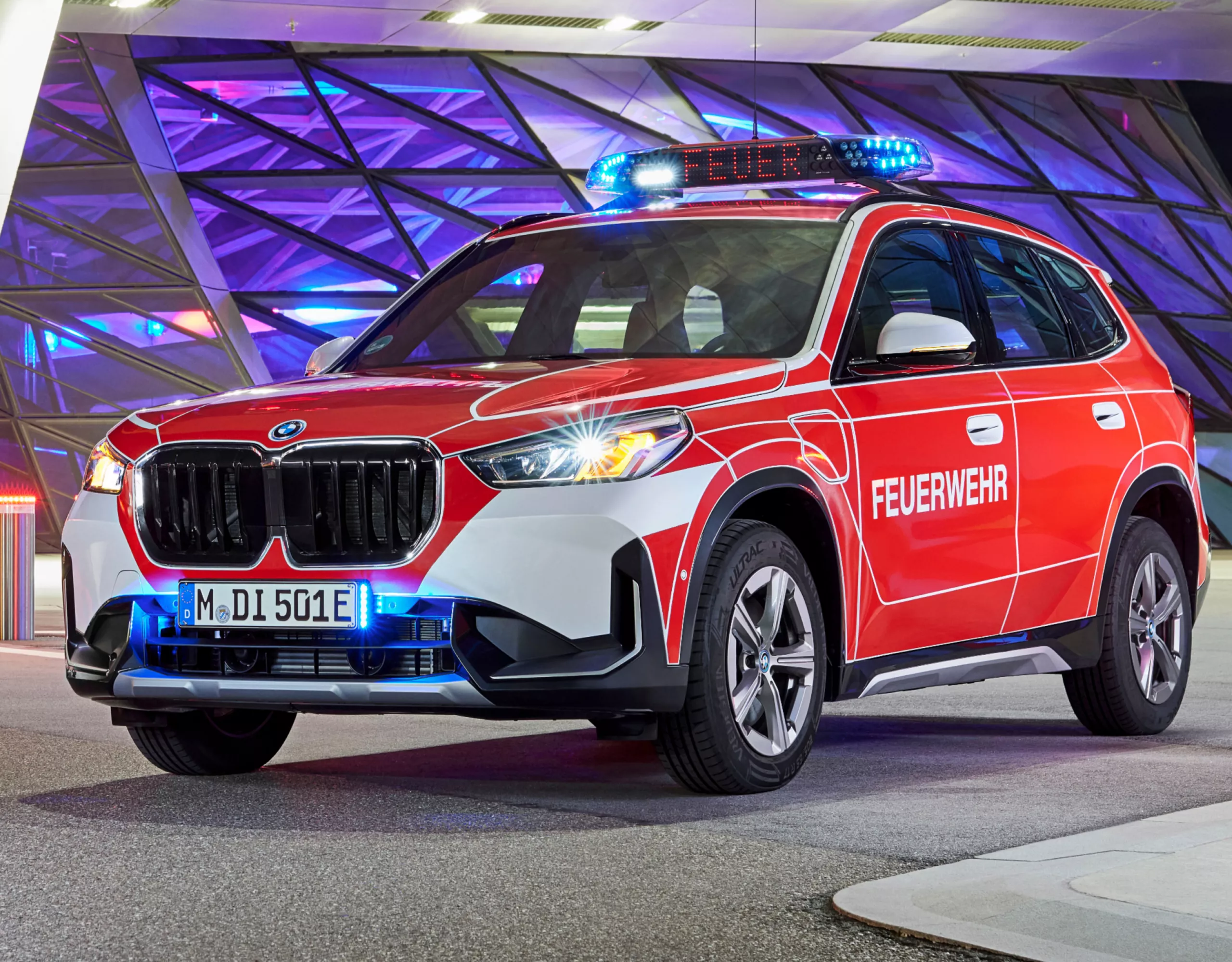 Voor de ingang van een modern gebouw, verlicht in roze en paars, staat een BMW brandweerauto