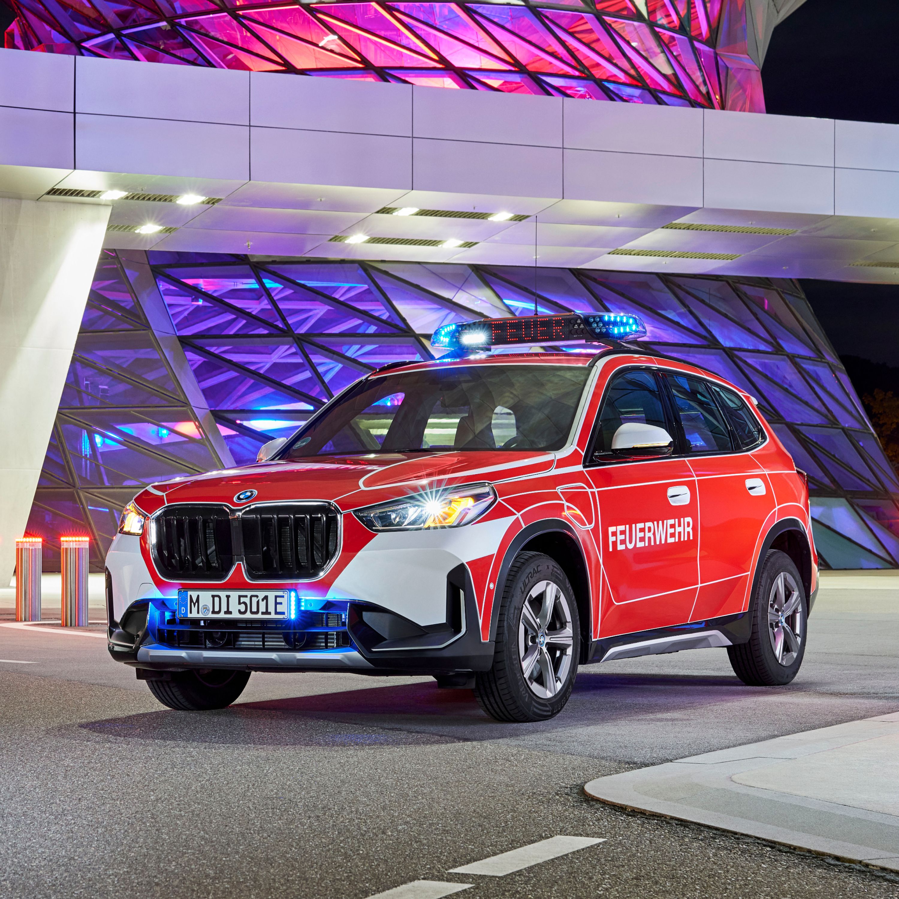 A BMW fire brigade vehicle parked in front of a modern building illuminated in pink and purple