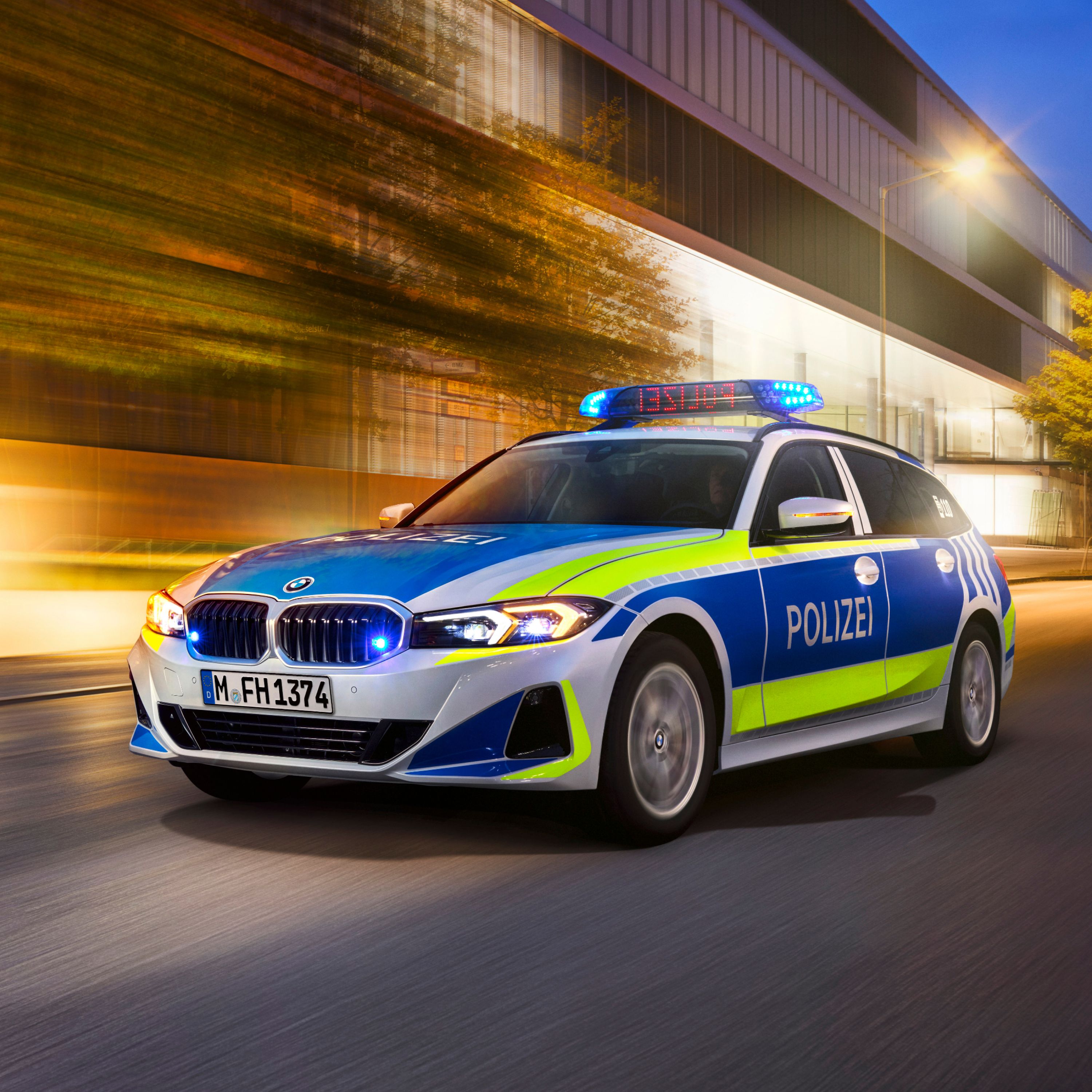 A BMW police vehicle drives past a lit-up building by twilight