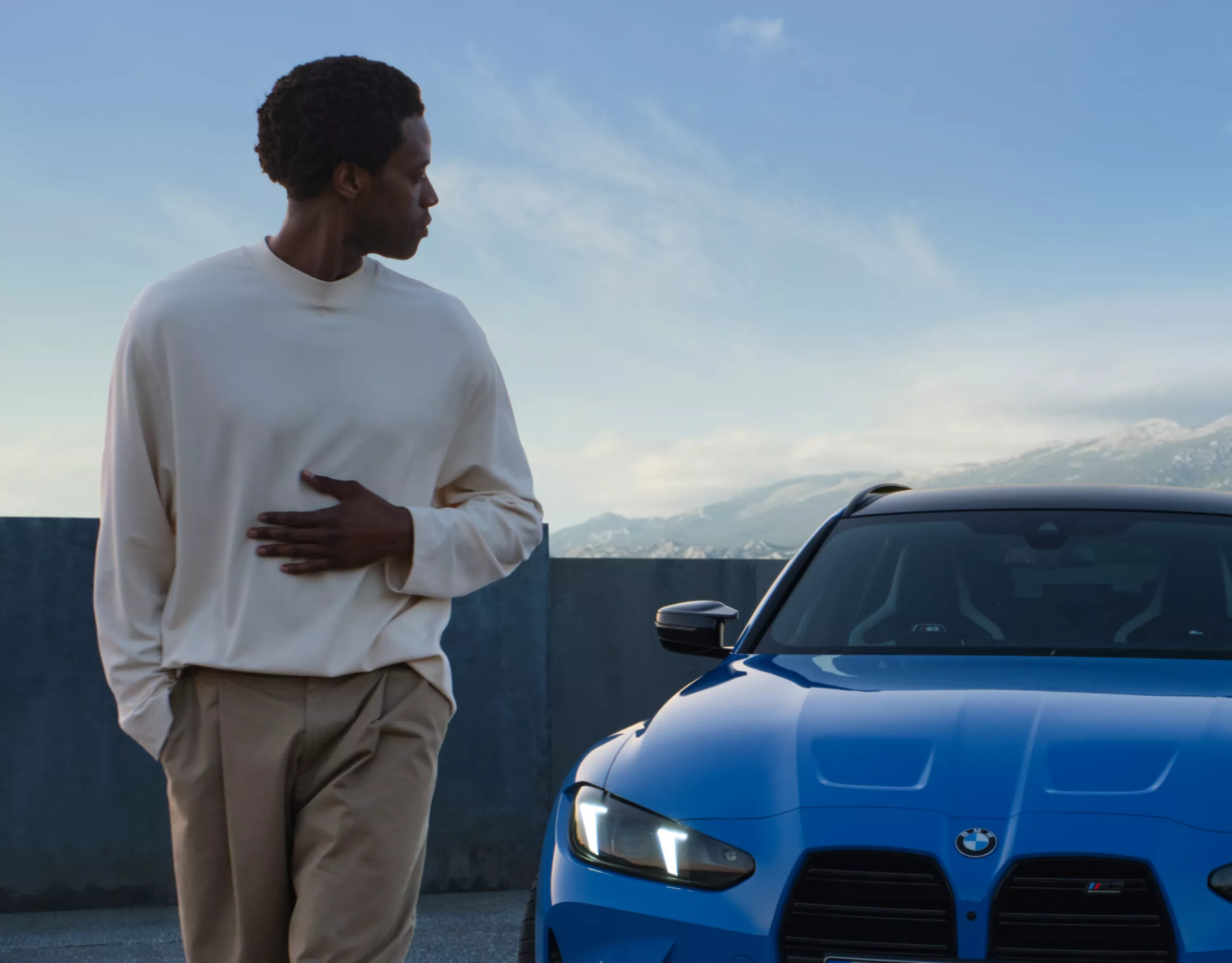 A man looks back at a BMW in the evening light with mountains in the distance