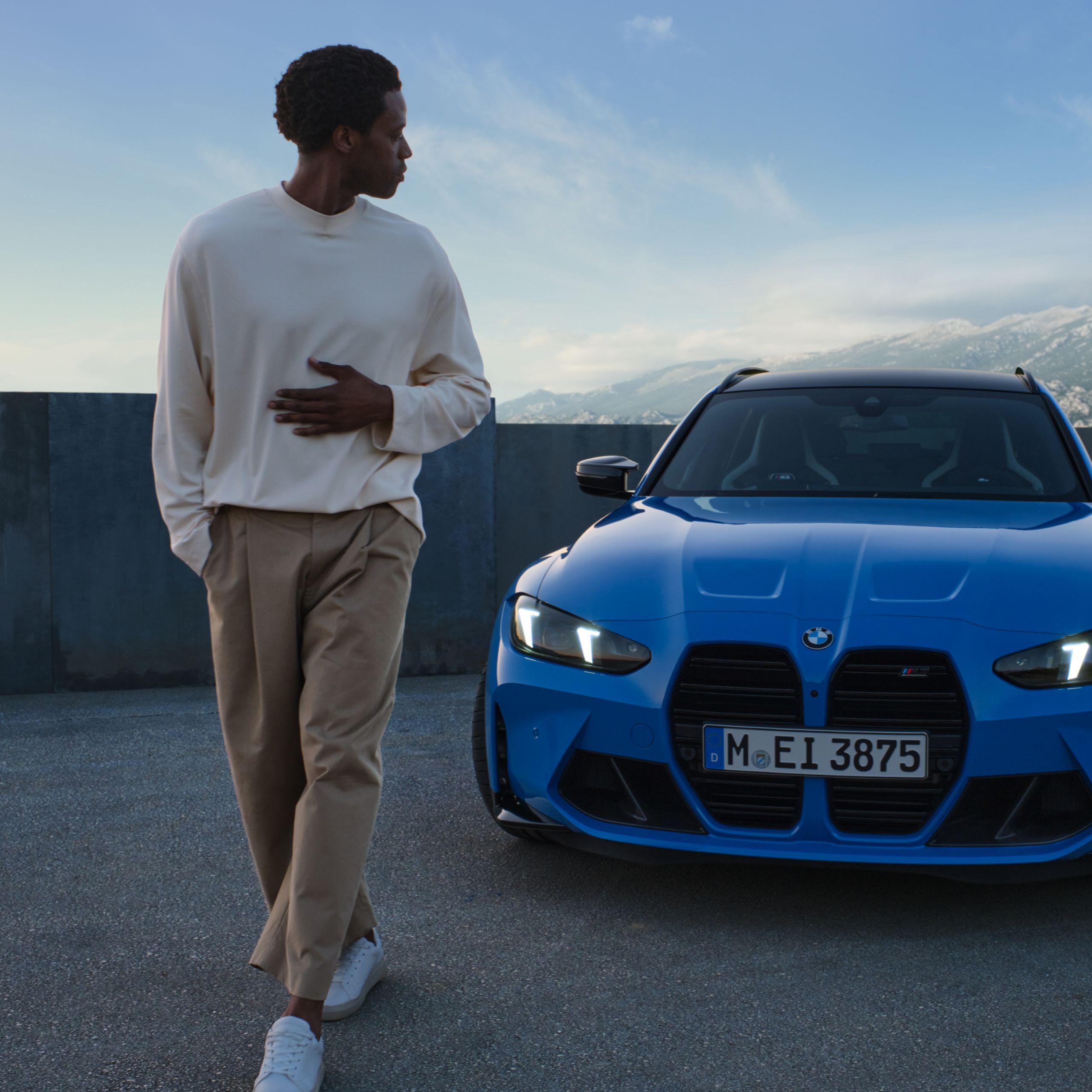 Un hombre mirando hacia atrás a un BMW al atardecer con montañas en la distancia