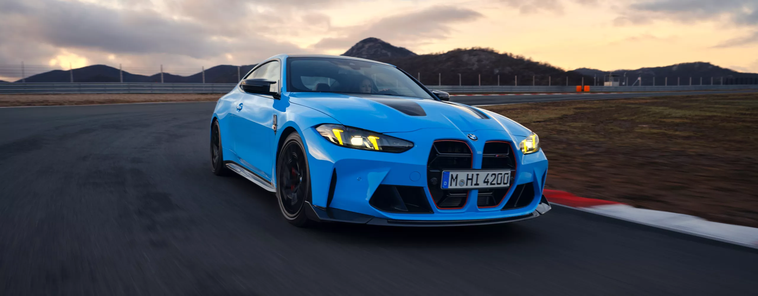 Sportowe, niebieskie BMW M4 na torze wyścigowym na tle zachodzącego słońca.