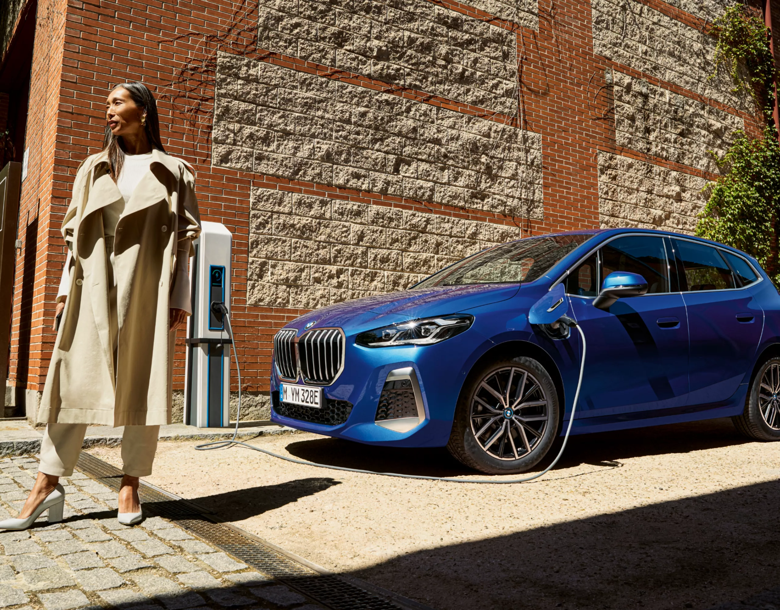 Egy személy elsétál egy téglafalra szerelt wallboxra csatlakoztatott BMW 2 Active Tourertől