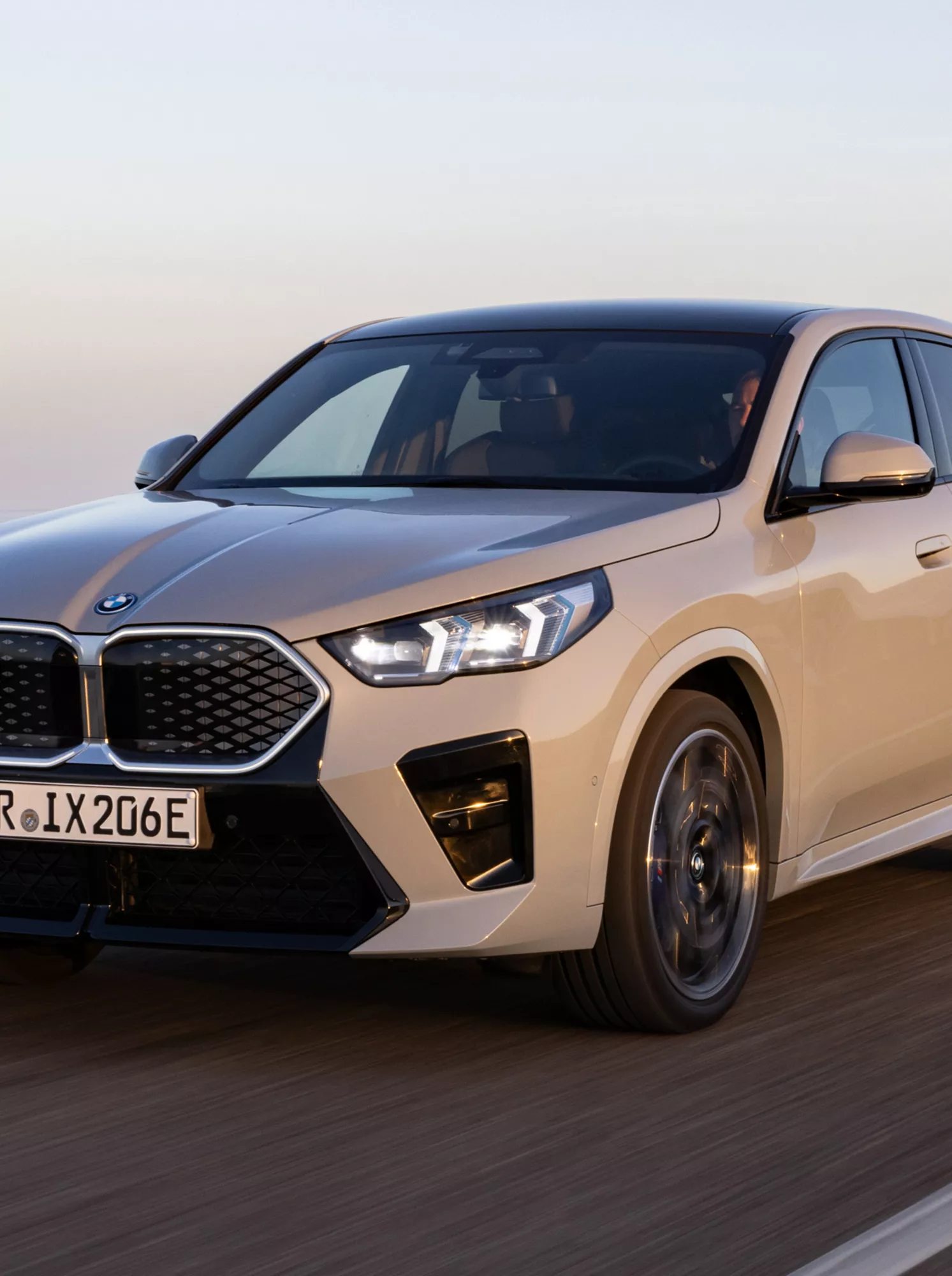BMW iX2 Elektroauto in Brooklyn Grau, unterwegs auf einer Straße bei Sonnenuntergang mit Blick aufs Meer im Hintergrund