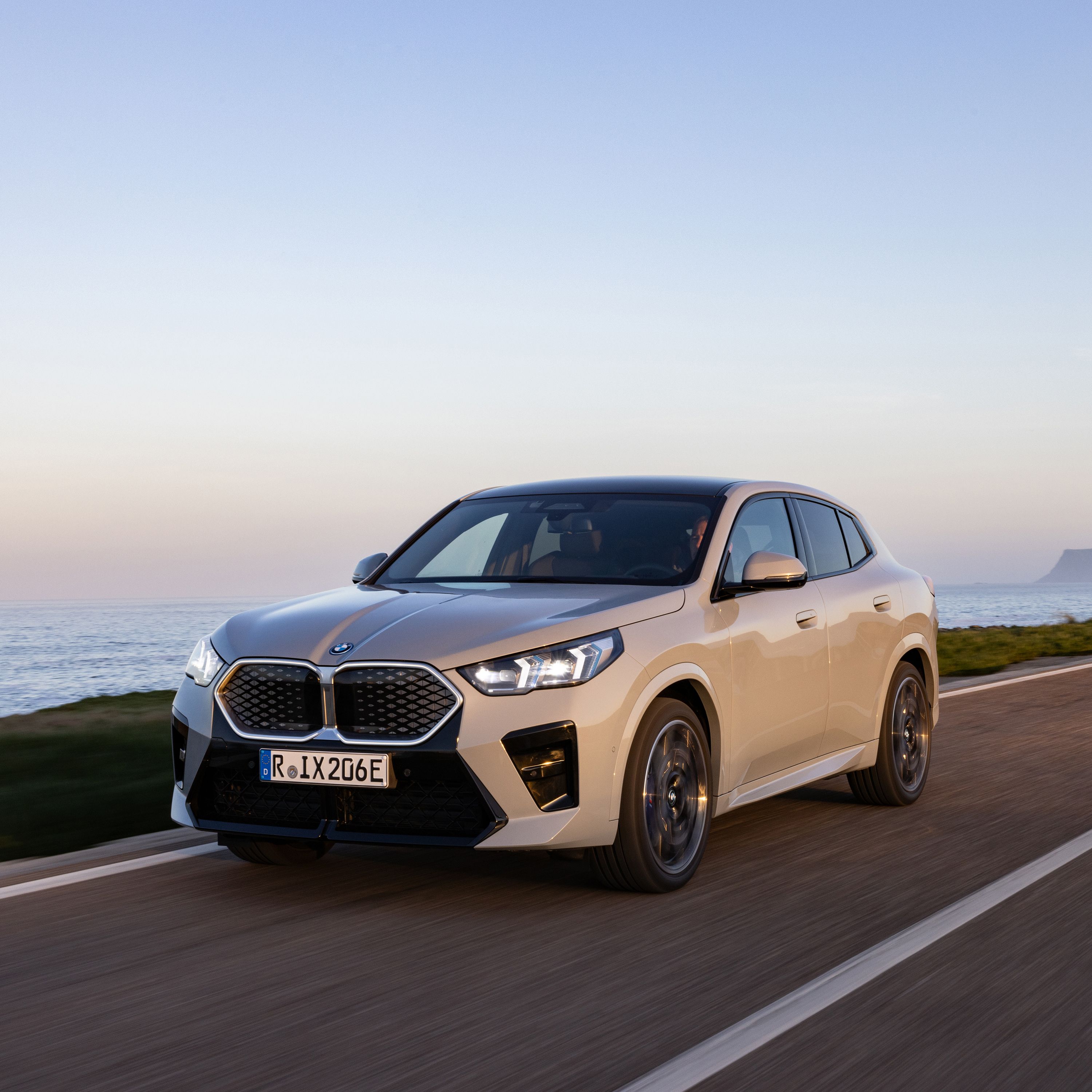 BMW iX2 all-electric vehicle in Brooklyn Grey color driving on a road at sunset with an ocean view in the background.