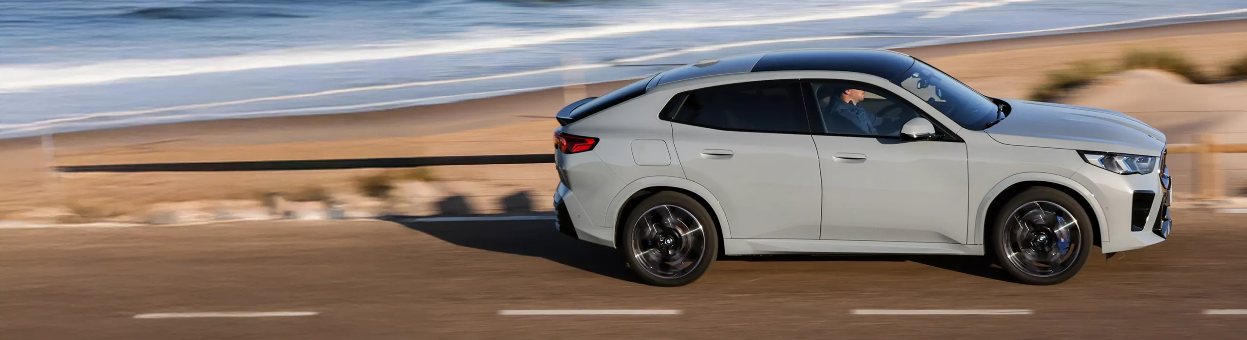 Egy M Brooklyn szürke metál színű BMW iX2 egy parti úton halad