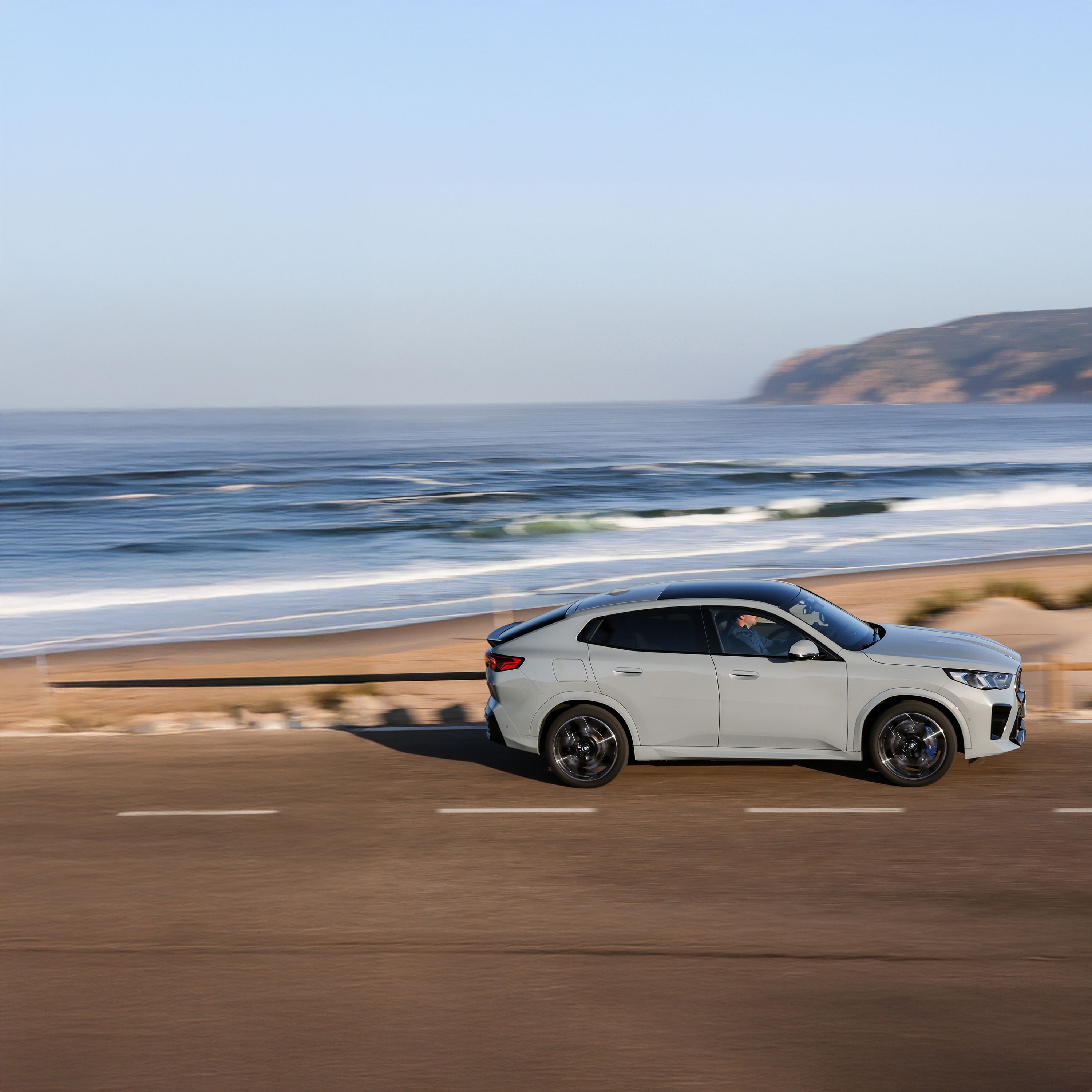 Una BMW iX2 elettrica di colore Brooklyn Grey che percorre una strada con vista sull’oceano e un cielo azzurro sullo sfondo