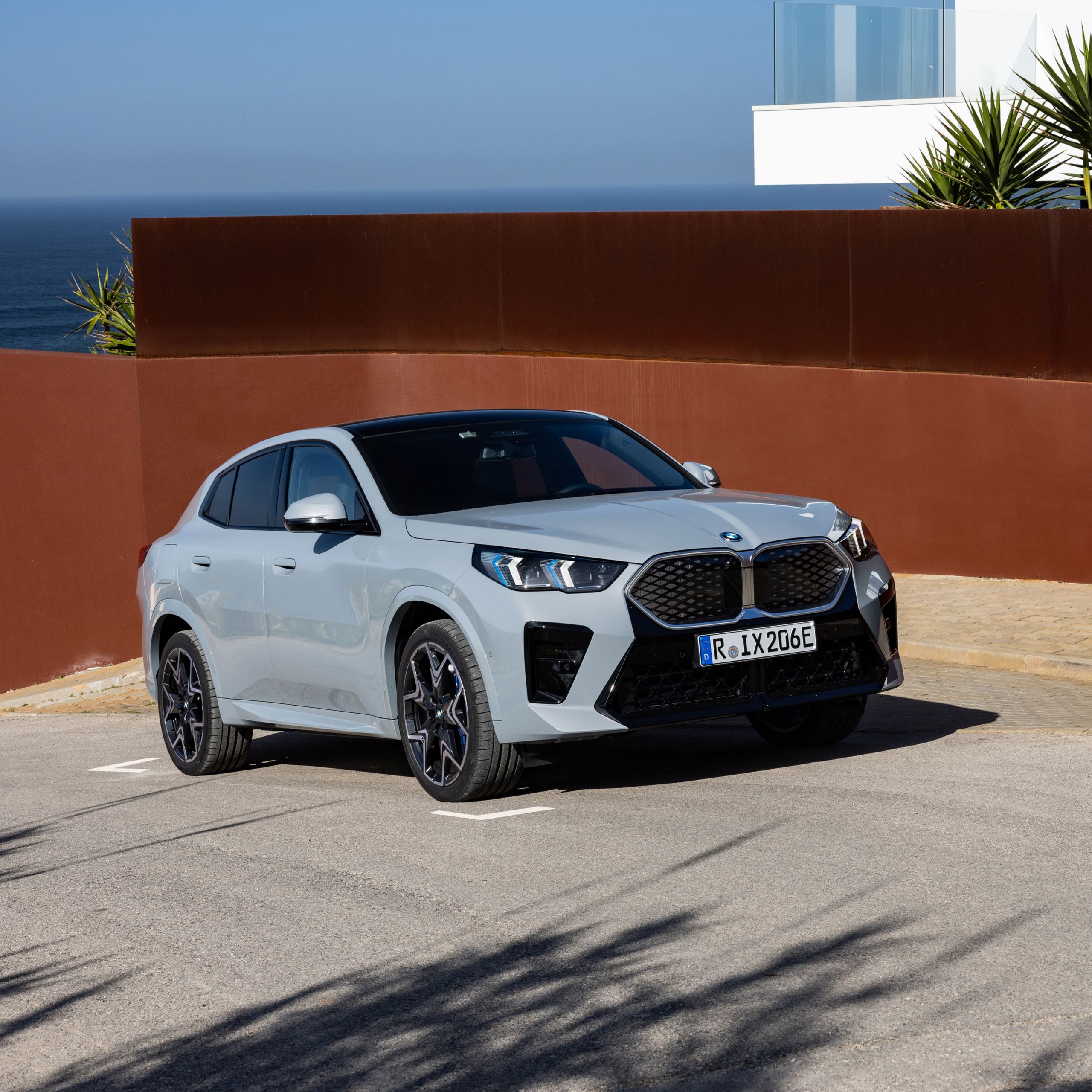 100% elektrické BMW iX2 v šedé barvě Brooklyn Grey zaparkované na silnici před domem s výhledem na oceán a modré nebe