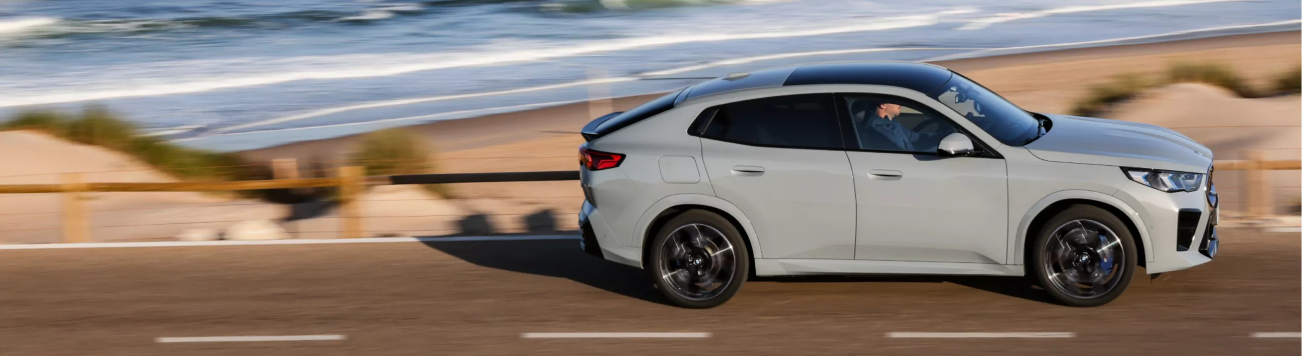 BMW iX2 Elektroauto in Brooklyn Grau, unterwegs auf einer Straße mit Blick aufs Meer und blauem Himmel im Hintergrund