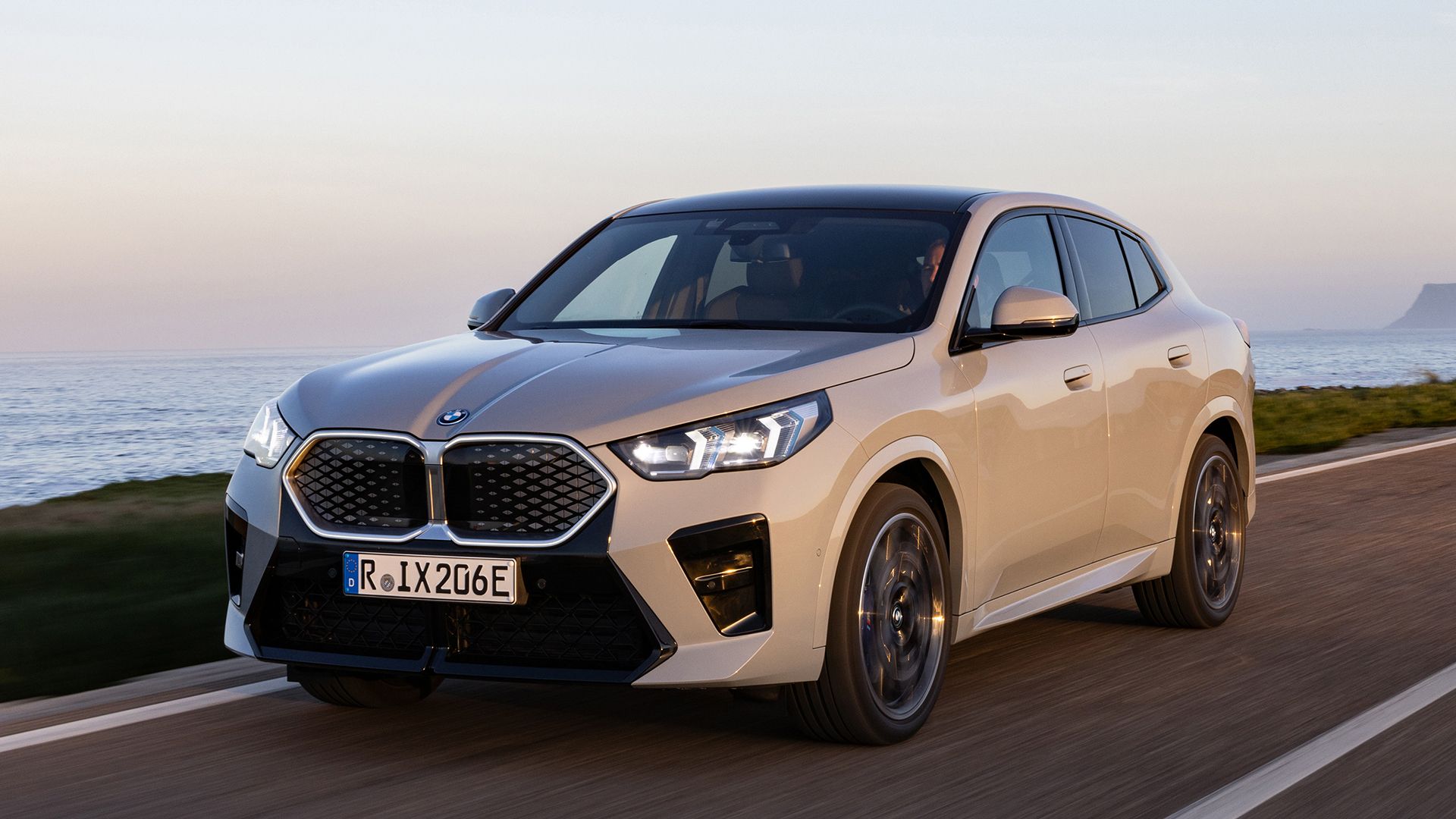  Elektrische BMW iX2 in de kleur Brooklyn Grey tijdens zonsondergang rijdend op een weg met de zee op de achtergrond.