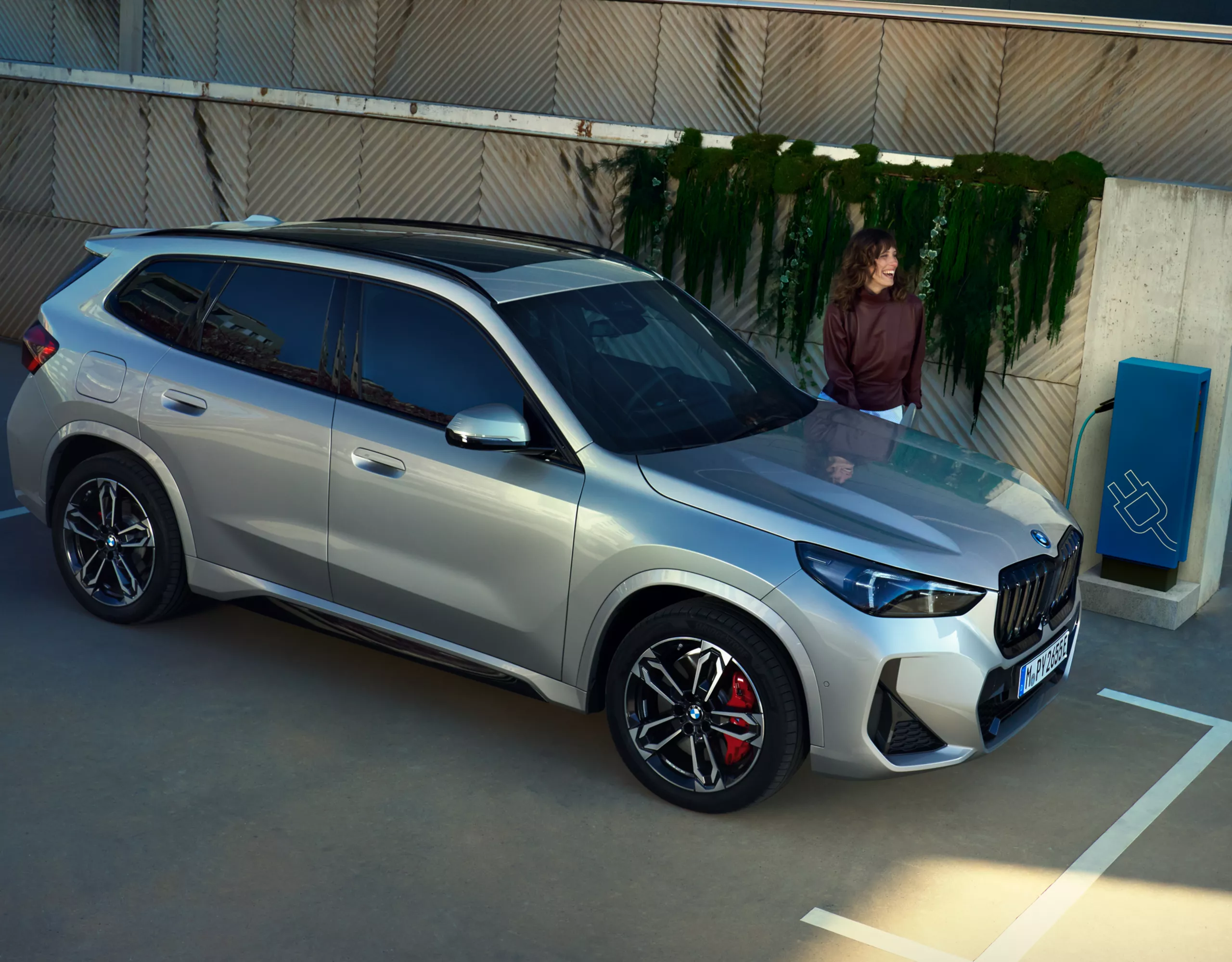 Ein BMW X1 Plug-in-Hybrid beim Aufladen an einer Ladestation. Eine Frau wartet in der Nähe.