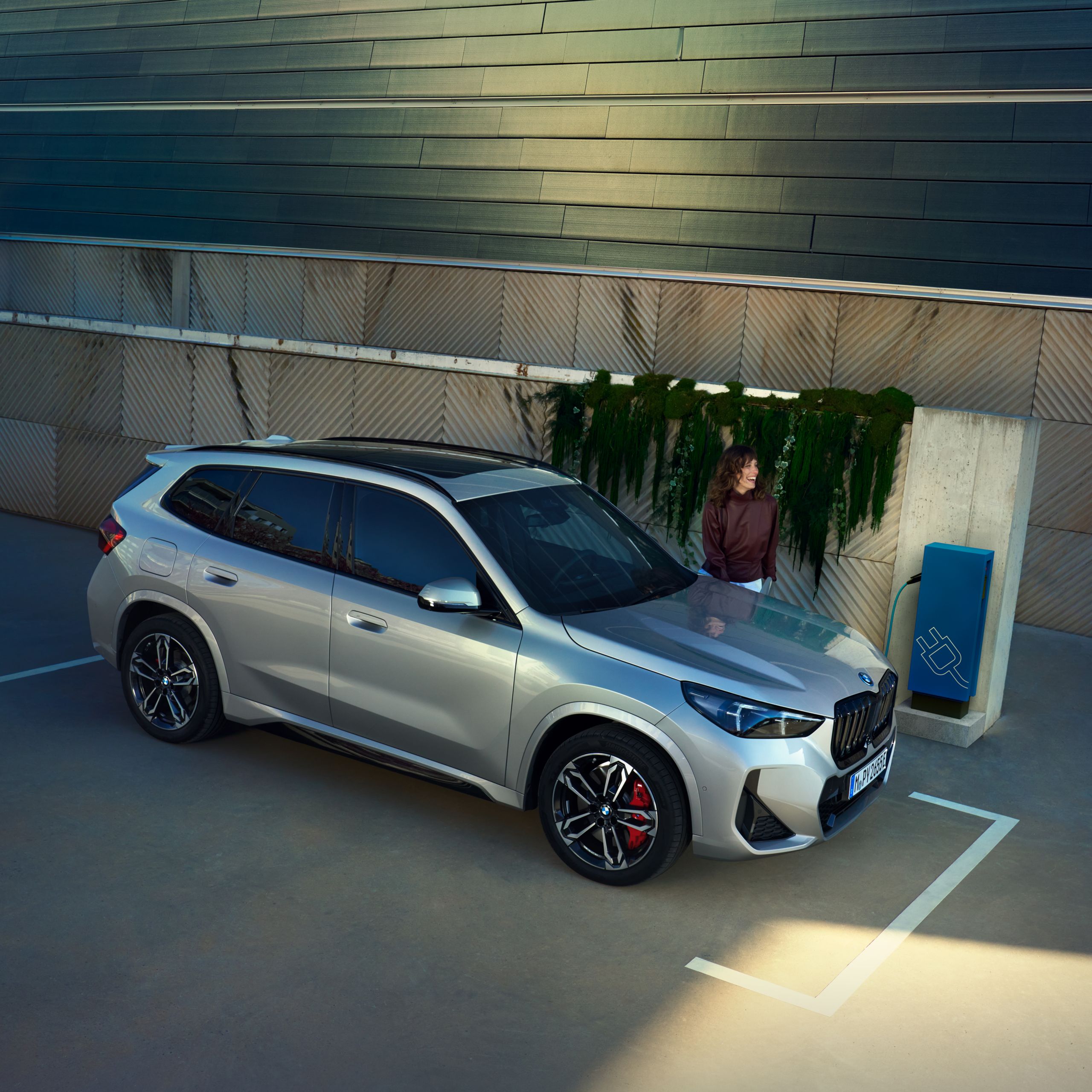 Ein BMW X1 Plug-in-Hybrid beim Aufladen an einer Ladestation. Eine Frau wartet in der Nähe.