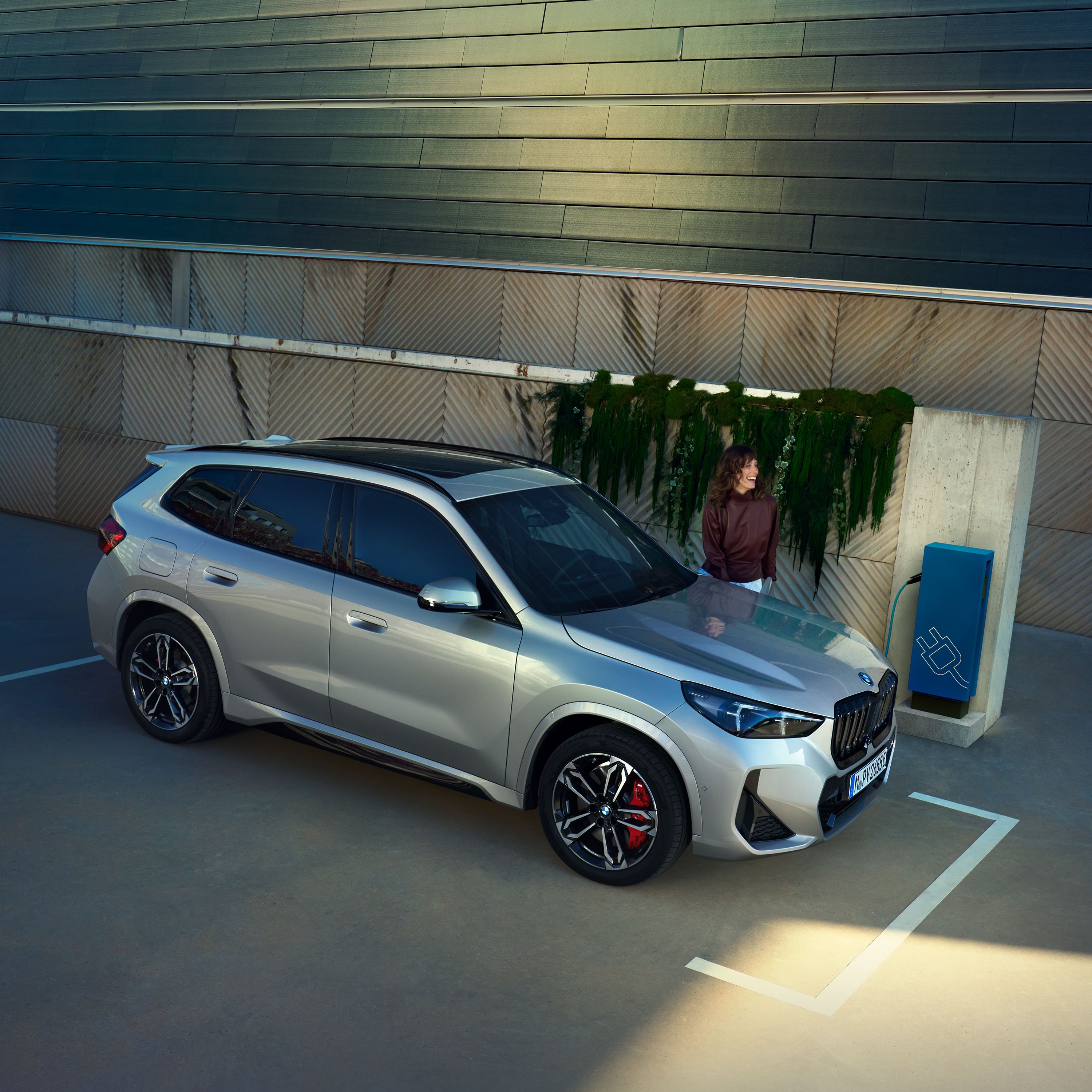 BMW X1 jako hybryda plug-in podczas ładowania na stacji ładowania. Kobieta czeka w pobliżu.