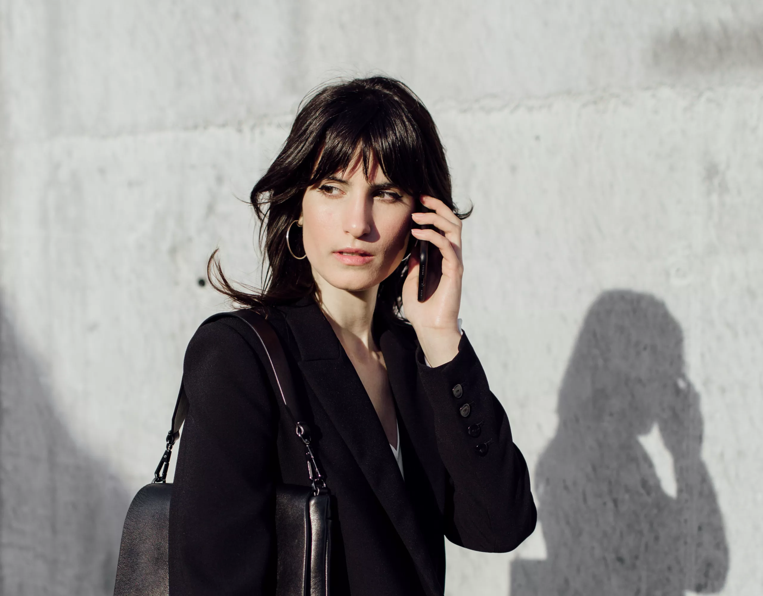 Eine Frau in dunkler Kleidung steht vor einer hellen Betonwand und hält ein Smartphone am Ohr