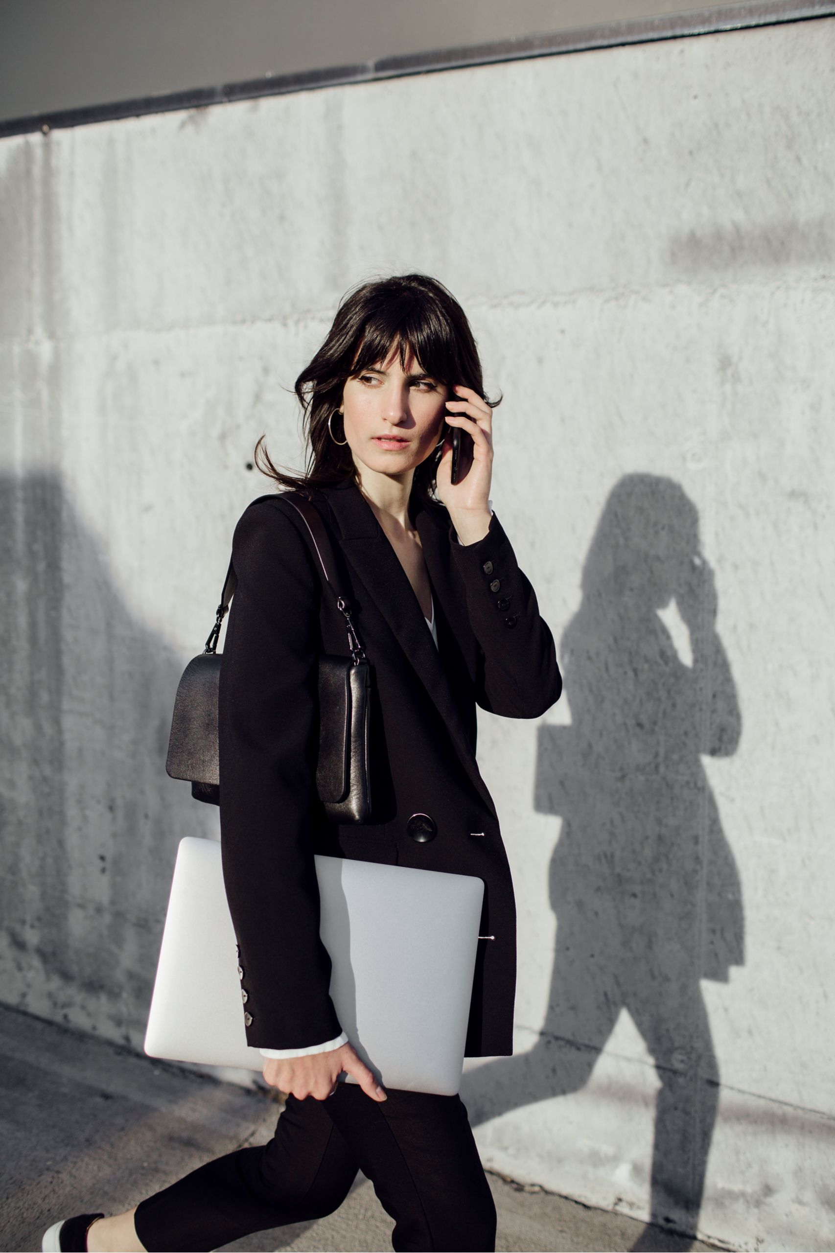Eine Frau in dunkler Kleidung steht vor einer hellen Betonwand und hält ein Smartphone am Ohr